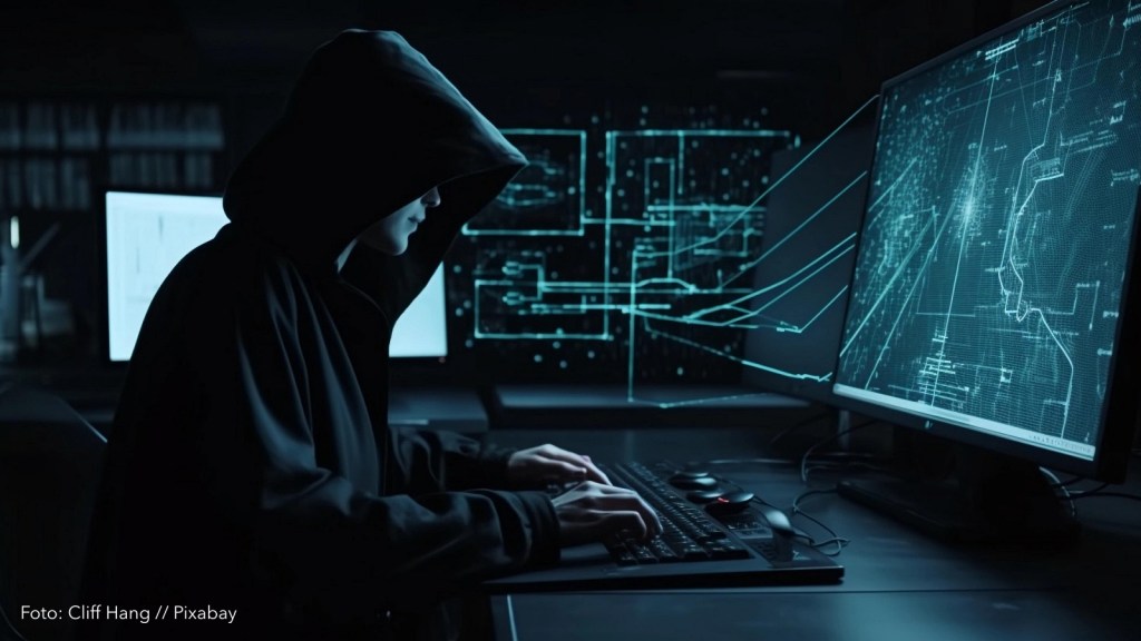 Eine Person mit dunklem Hoodie sitzt in einem dunklen Raum und macht verdächtige Dinge an einem Computer.